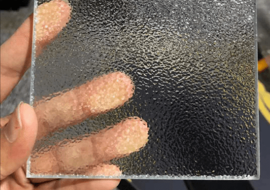 Hand holding a square glass pane with a frosted, textured surface, partially revealing the fingers behind it.