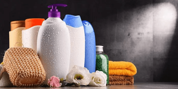 A collection of personal care products including bottles of shampoo, conditioner, body wash, and a loofah, towel, and bath salts, with a steamy background, suggestive of a spa or bathroom setting.