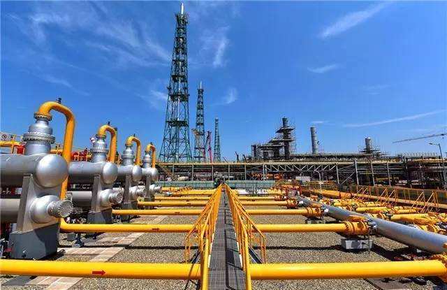 Complex network of yellow pipelines at a petrochemical plant with a distillation tower in the background, related to Propylene Carbonate applications in gas treatment.