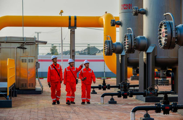 Three workers in red safety suits inspecting the natural gas pipelines and valves at an industrial facility.