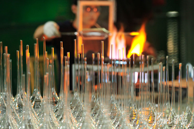 Artisan glassblowing process with clear glass rods in focus and a craftsman working with a blazing furnace in the background.