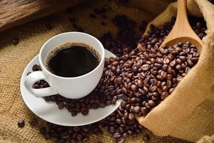 Cup of black coffee surrounded by roasted coffee beans and a burlap sack, representing the ethyl acetate decaffeination process.